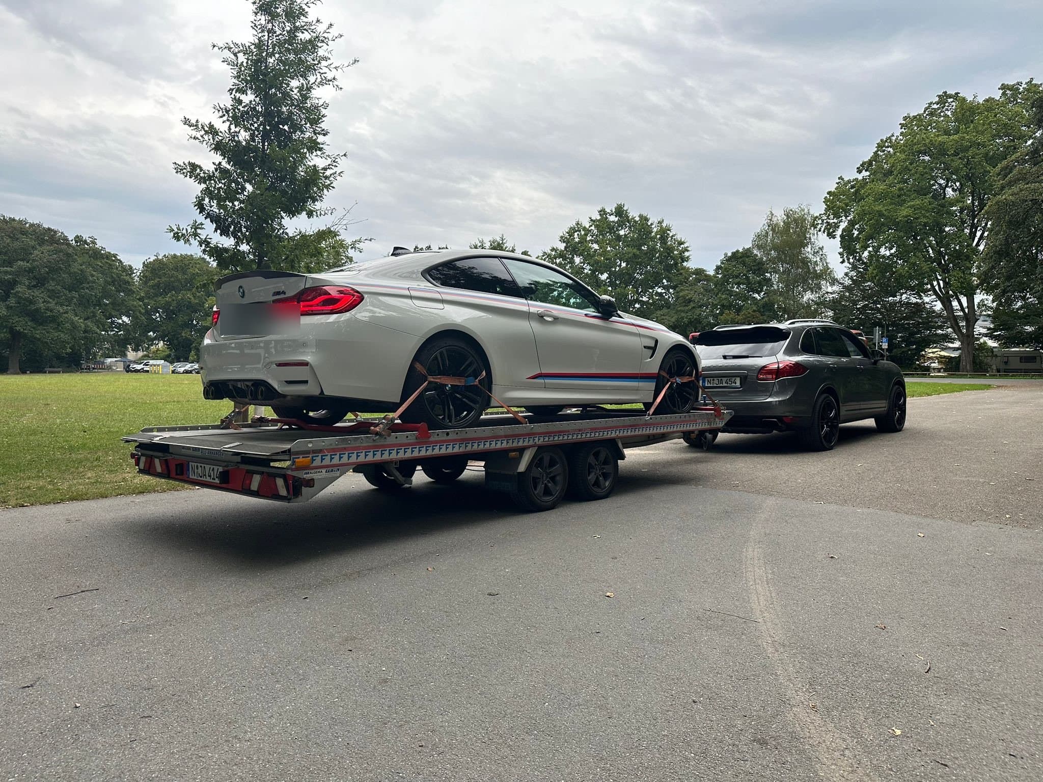 Weißer BMW M4 auf offenem Autotransportanhänger hinter grauem Porsche Cayenne